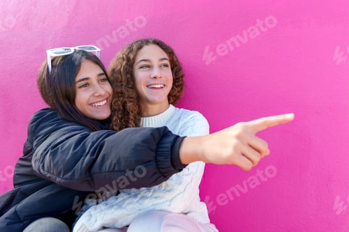 Preview: Cheerful female friends embracing, playfully pointing fingers near bright pink wall
