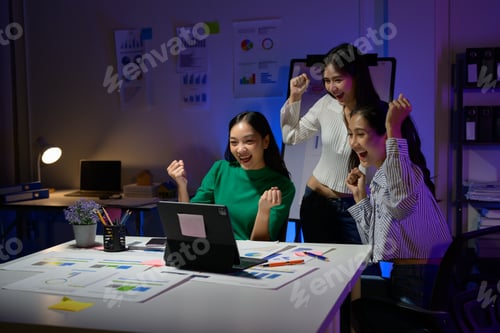 Preview: Excited Team Celebrating Success in Modern Office with Charts and Laptop at Night