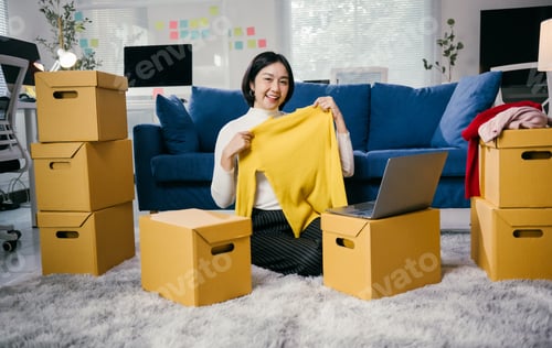 Preview: Asian female entrepreneur showing clothes in a live stream using laptop at home office