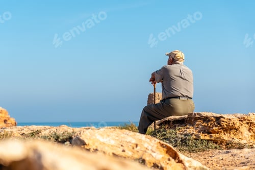 Preview: Pensive old man in beret sitting and with walking stick on the shore by the sea