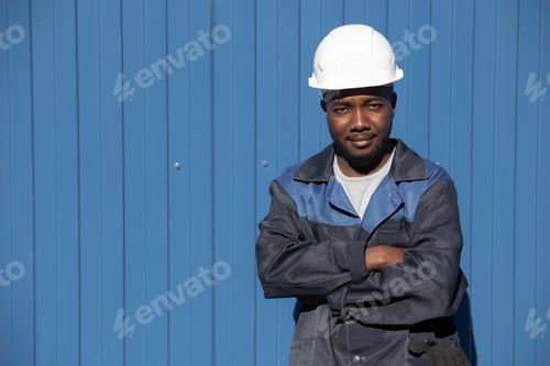 Preview: Happy young factory worker of African ethnicity in workwear and hardhat crossing his arms by chest