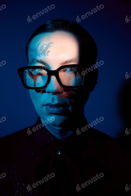 Preview: Stylish young man with striking blue tones and unique glasses poses confidently in artistic lighting