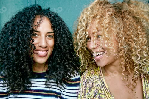 Preview: Portrait of two young women with long curly black and blond hair, smiling at camera.