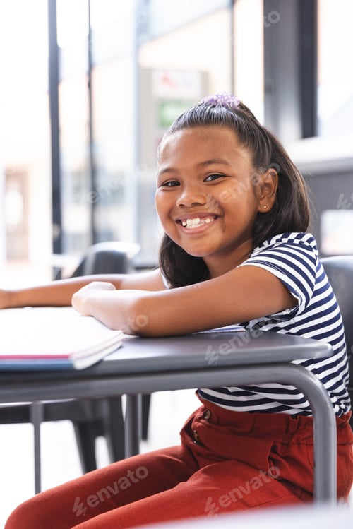 Preview: In school, young biracial girl with a bright smile sitting at a desk in a classroom