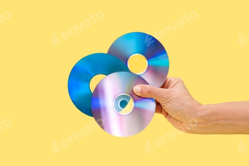 Preview: Close up of Hand holding three colorful cds on yellow background