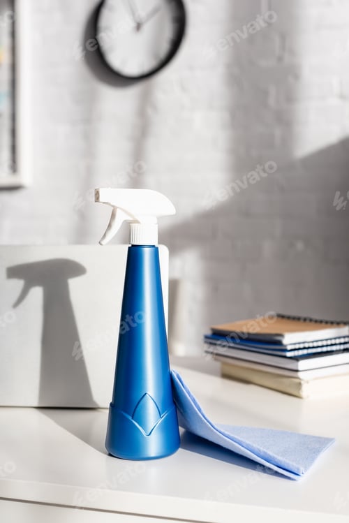 Preview: Bottle of detergent with sprayer and rag near laptop and notebooks on blurred background on table