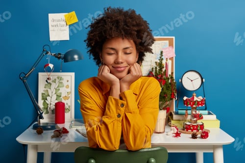 Preview: Pleased dark skinned woman keeps hands under chin, sits at comfortable chair with eyes closed, poses
