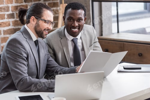 Preview: handsome businessman reading document while african american partner looking at camera in office