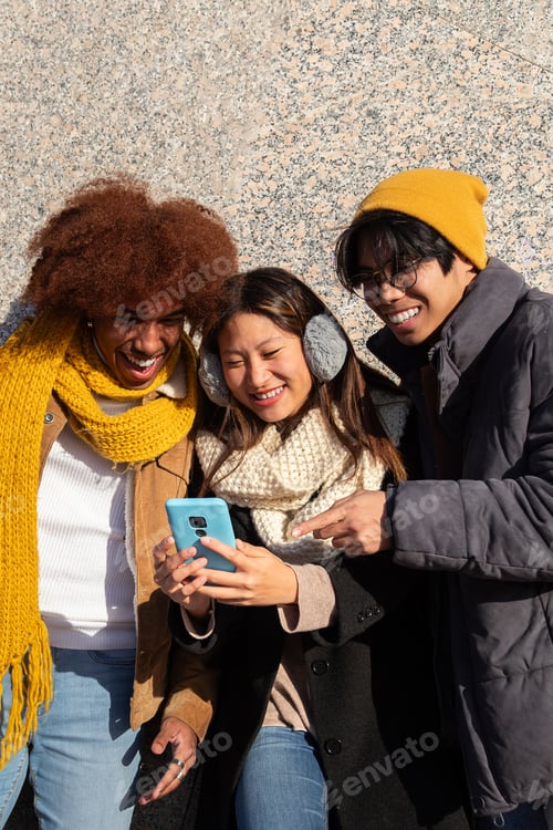 Preview: Happy university student friends having fun using phone together during sunny winter day. Vertical