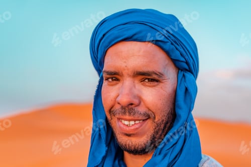 Preview: Close-up of Berber man in traditional blue turban in Sahara desert at sunset smiling at camera