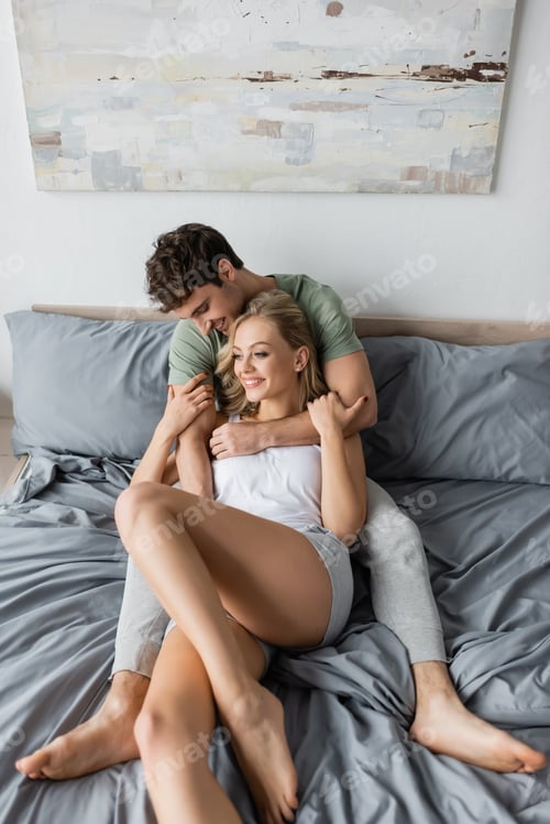 Preview: Positive man in pajama hugging blonde girlfriend on bed in morning