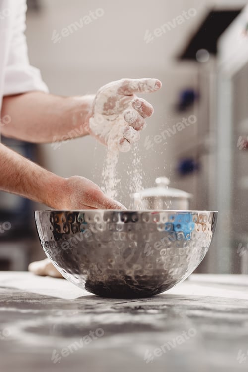 Preview: Baker adds flour to dough in an iron bowl on the table in the bakery