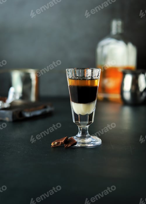 Preview: A three-layer cocktail shot with whiskey and liqueurs in a glass and chocolate next to it on a dark