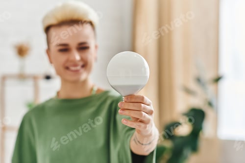 Preview: energy saving light bulb in hand of overjoyed young woman with trendy hairstyle standing at home