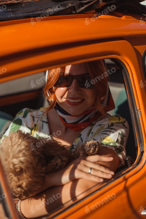 Preview: A young woman in a silk headscarf sits in a small orange retro car and plays with a small dog
