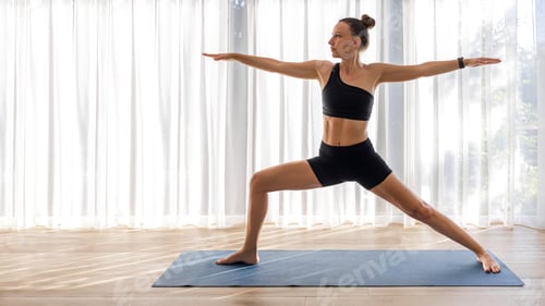 Preview: Yoga woman in black sportswear standing in warrior pose on the mat in big room