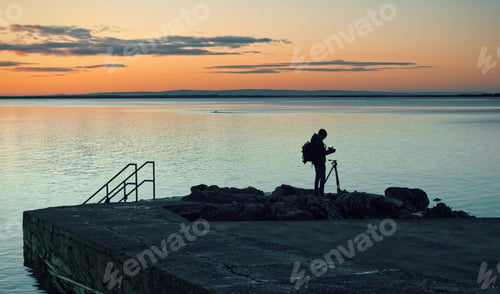 Preview: Silhouetted photographer taking shot of Blacrock tower at sunrise on Salthill beach in Galway, Irela