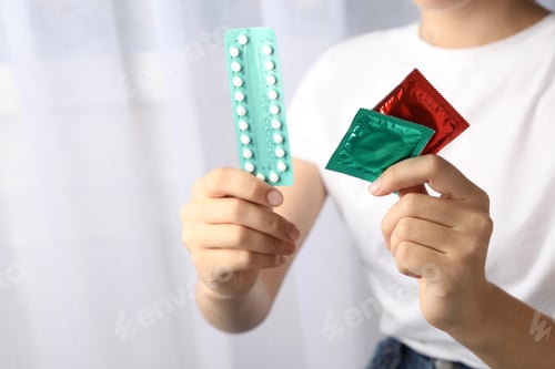 Preview: Young woman holding birth control pills and condoms on light background, closeup. Safe sex concept
