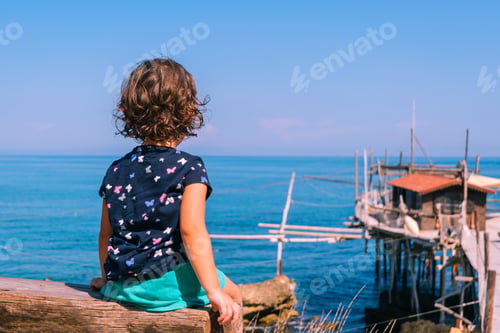 Preview: A girl with curly hair sits on a bench overlooking the sea and an old trabocco.