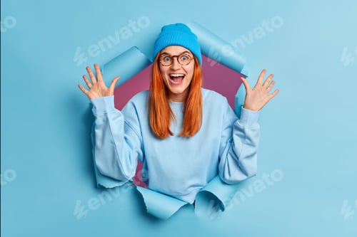 Preview: Glad redhead young woman wears optical glasses hat and jumper raises hands stands in torn paper hole