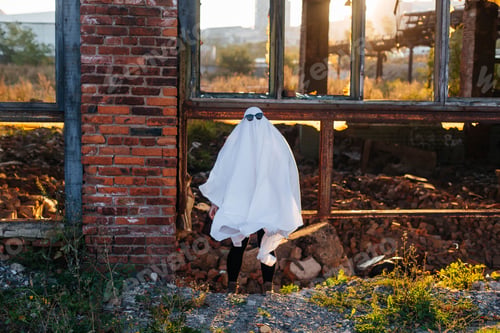 Preview: Girl in white sheet and sunglasses in a ghost costume on Halloween walks near a destroyed building
