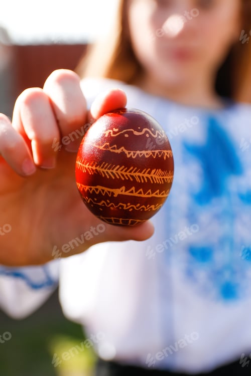 Preview: Defocus ukrainian girl in vyshyvanka holding one colored red egg on nature background. Easter, Ukrai