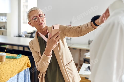 Preview: Mature clothes designer takes photo of new dress model with mobile phone in sewing workshop