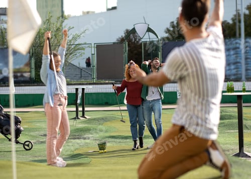 Preview: Group of smiling friends enjoying together playing mini golf in the city.