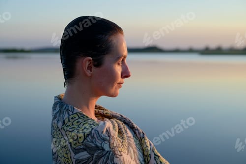 Preview: A woman stands by a tranquil body of water at dusk, her profile captured against a soft sunset sky.
