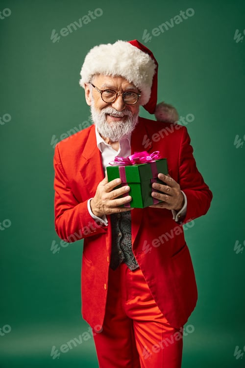 Preview: stylish Santa with beard and christmassy hat holding present in front of camera, winter concept