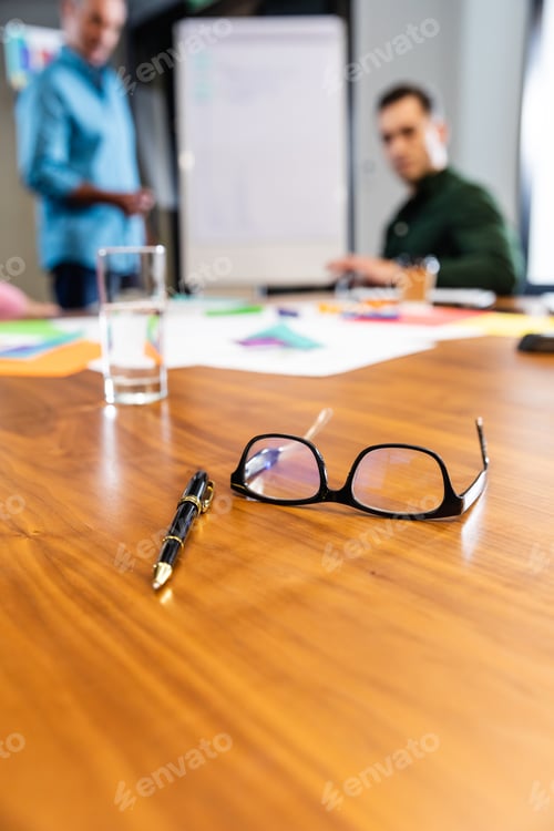 Preview: Pen and eyeglasses on conference table while business colleagues in background at office