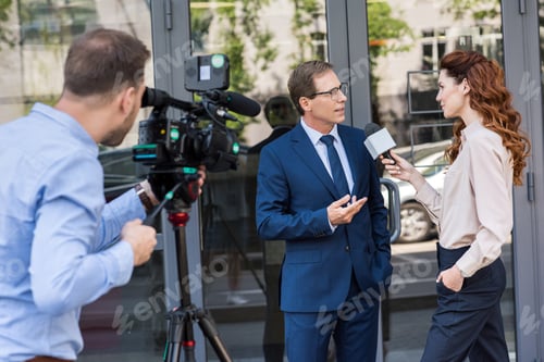 Preview: cameraman and news reporter with microphone interviewing businessman near office building