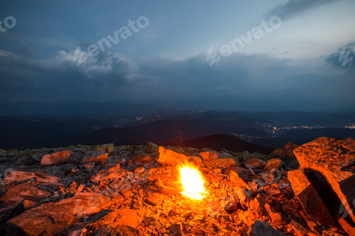 Preview: Beautiful summer night in mountain. Bright fire burns on rocky mountain top under blue cloudy sky.