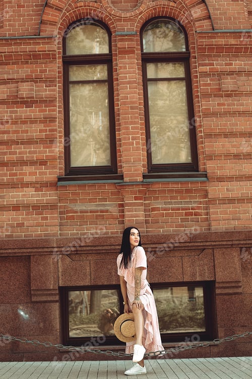 Preview: A young brunette in a soft pink dress with a hat walks around the city