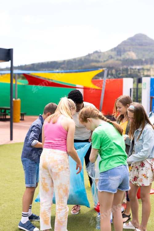 Preview: Vertical of diverse female teacher and pupils collecting recycling in schoolyard, with copy space