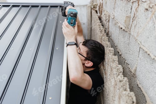 Preview: A young guy with a drill is installing a roof on a metal barn.
