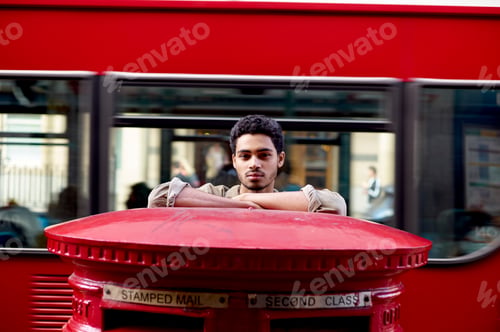 Preview: Man leaning on a red Royal Mail postbox with a blurred double-decker bus in the background.