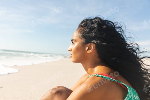 Preview: Side view of thoughtful young biracial woman with long black hair looking away at beach