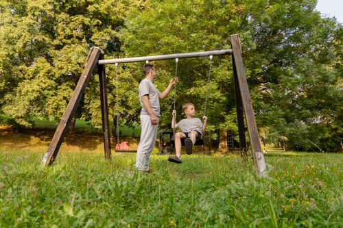 Preview: A father pushes his young son on a swing at a playground located in a green area.