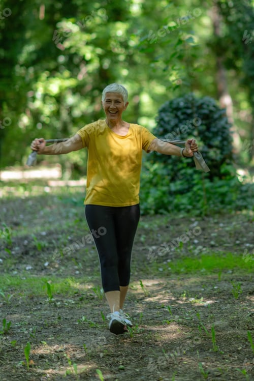 Preview: Active senior woman exercising with elastic resistance band for strength training in nature.