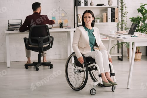 Preview: attractive disabled businesswoman sitting in wheelchair in office near colleague