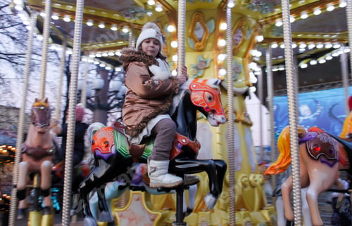 Preview: Funny toddler girl in warm clothes rides a merry-go-round horse during the winter Christmas fair
