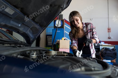 Preview: Young casual woman in shirt and gloves working with car in repair service checking engine oil level