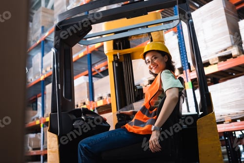 Preview: happy female forklift operator in hard hat and safety vest looking at camera in warehouse, cargo