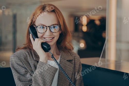 Preview: Female office worker in spectacles talking on landline phone with co-worker and smiling at camera