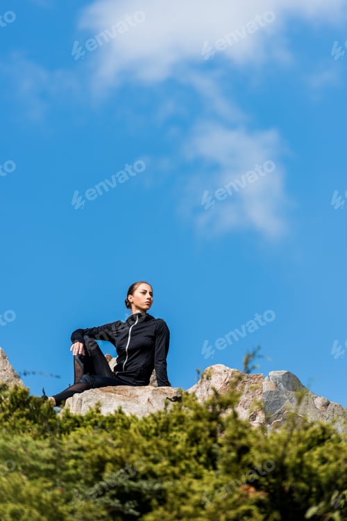 Preview: young woman sitting on rocks and looking away in front of blue sky