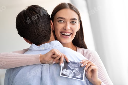 Preview: Happy european young wife hugs husband and holds ultrasound picture of baby in living room interior