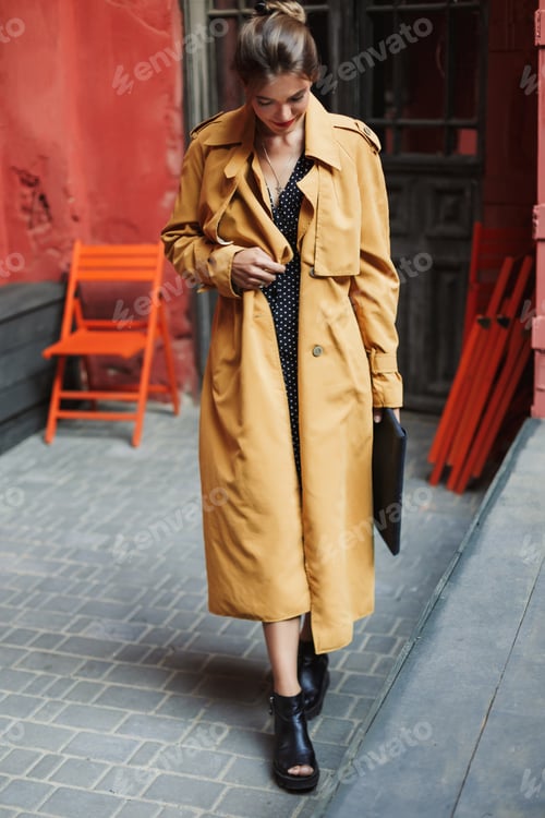 Preview: Young beautiful smiling woman in trench coat and boots holding b