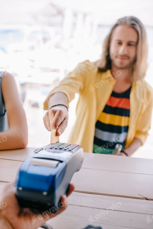 Preview: selective focus of young man paying with credit card on terminal at beach bar