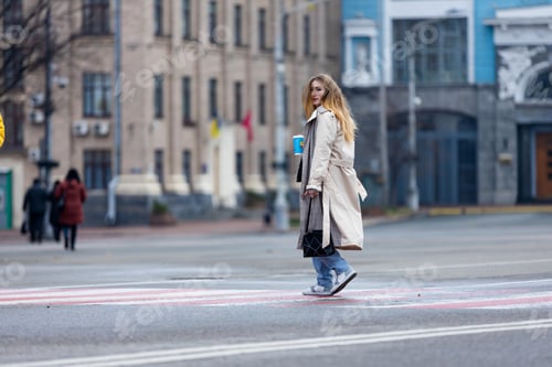 Preview: Shot of a pretty young woman in coat crossing the street holding coffee cup and looking away.